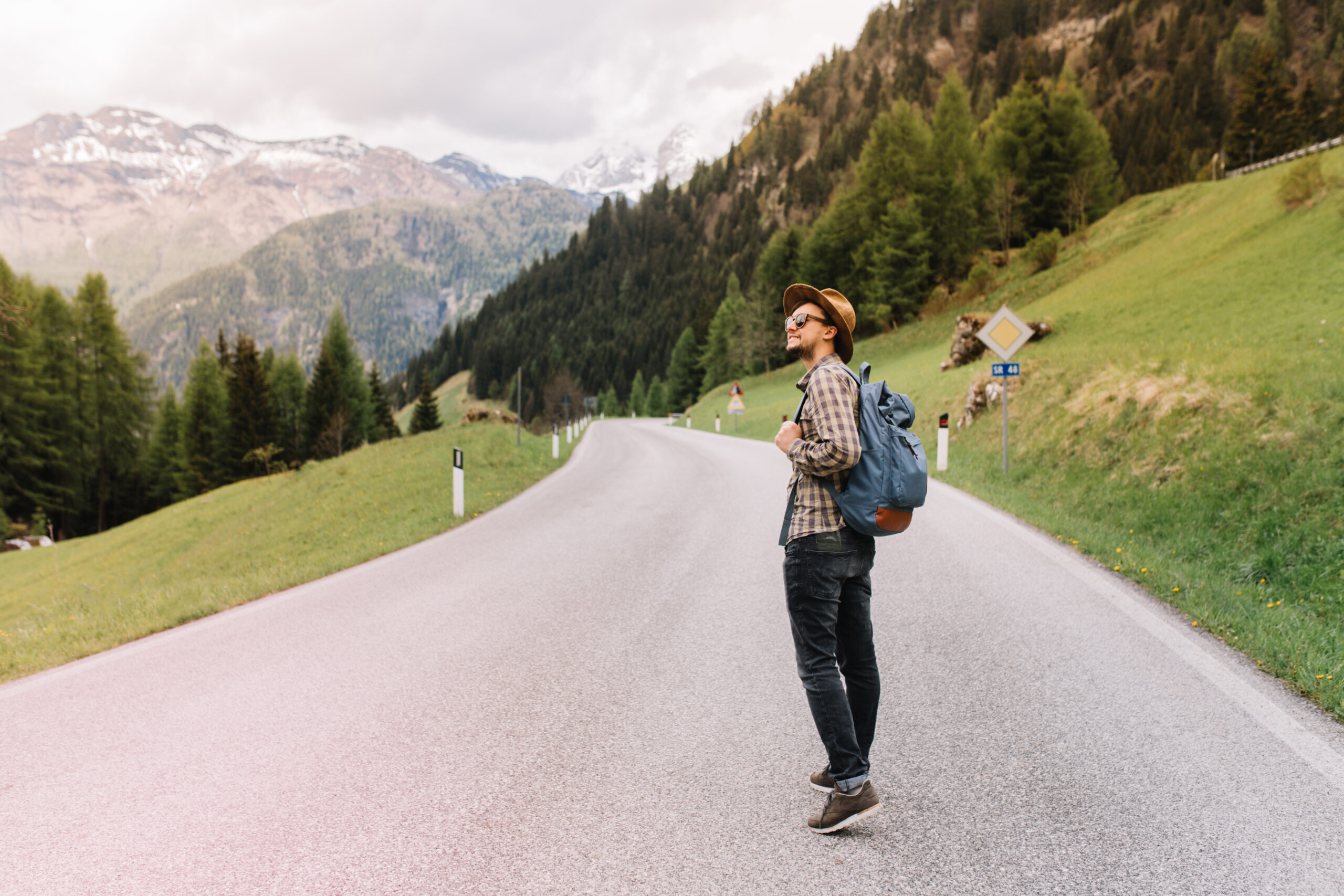 A young man on a solo mountain tour, standing on a road with the Alps in the background.