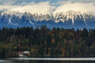 Scenic lake view with autumn forest, snowy mountains, and cozy houses – ideal for a travel agency promotion.