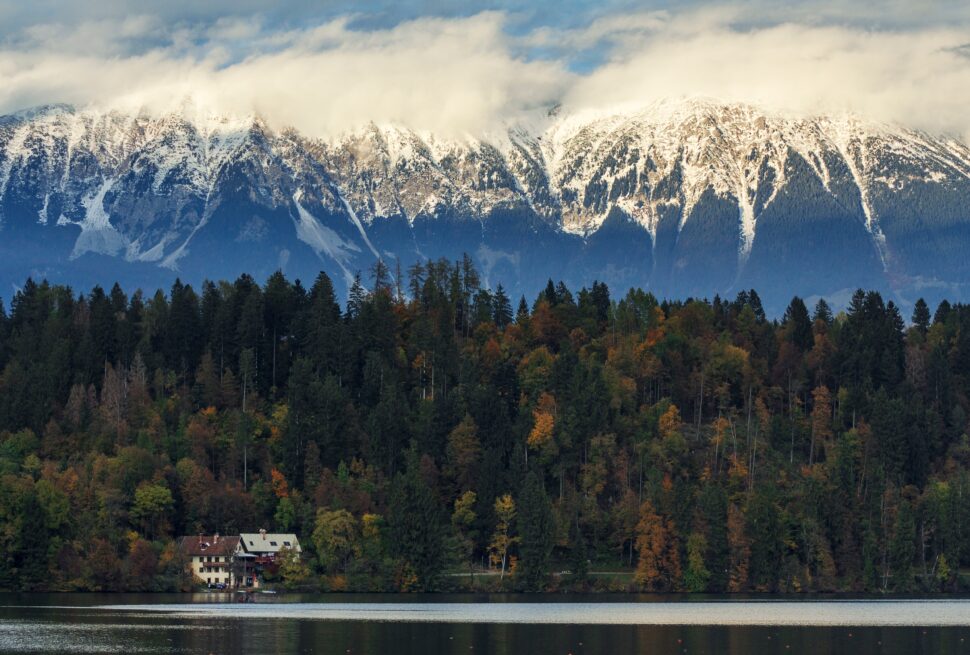 Scenic lake view with autumn forest, snowy mountains, and cozy houses – ideal for a travel agency promotion.